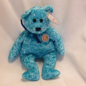 Puede incluir: Un oso de peluche turquesa con ojos negros y nariz negra. El oso tiene una cinta azul alrededor del cuello y una etiqueta circular en el pecho. Una etiqueta blanca est&aacute; sujeta a su oreja.