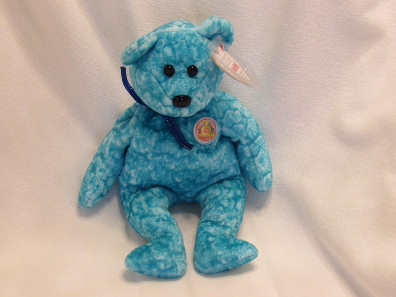 Puede incluir: Un oso de peluche turquesa con ojos negros y nariz negra. El oso tiene una cinta azul alrededor del cuello y una etiqueta circular en el pecho. Una etiqueta blanca est&aacute; sujeta a su oreja.