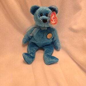 Puede incluir: Un oso de peluche azul con una cinta azul claro alrededor del cuello. El oso tiene una nariz negra y una etiqueta roja sujeta a su oreja. El oso tiene un n&uacute;mero 1 dorado en el pecho. El oso mide aproximadamente 20 cm de alto.