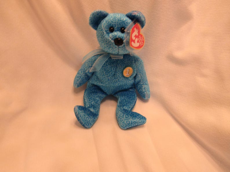 Puede incluir: Un oso de peluche azul con una cinta azul claro alrededor del cuello. El oso tiene una nariz negra y una etiqueta roja sujeta a su oreja. El oso tiene un n&uacute;mero 1 dorado en el pecho. El oso mide aproximadamente 20 cm de alto.