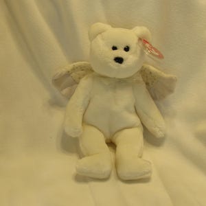 Puede incluir: Un oso de peluche blanco con alas de &aacute;ngel. El oso tiene una nariz negra y ojos negros peque&ntilde;os. Las alas son de color beige claro con un estampado de puntos. Una etiqueta roja est&aacute; adherida a la oreja del oso.