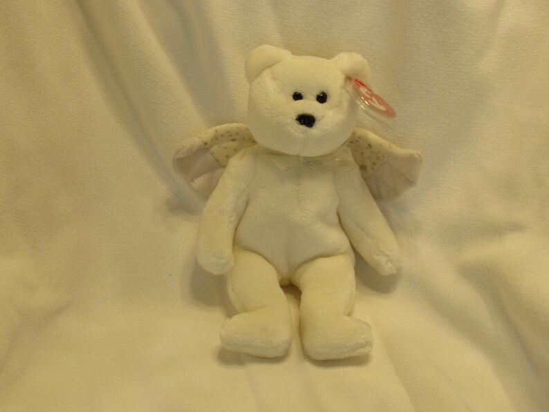 Puede incluir: Un oso de peluche blanco con alas de &aacute;ngel. El oso tiene una nariz negra y ojos negros peque&ntilde;os. Las alas son de color beige claro con un estampado de puntos. Una etiqueta roja est&aacute; adherida a la oreja del oso.