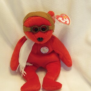 Puede incluir: Un oso de peluche rojo con gorro y gafas de aviador marrones, una bufanda blanca y un emblema circular. El oso tiene una nariz negra y una etiqueta Ty adjunta. El juguete es un art&iacute;culo de colecci&oacute;n.