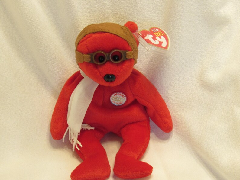 Puede incluir: Un oso de peluche rojo con gorro y gafas de aviador marrones, una bufanda blanca y un emblema circular. El oso tiene una nariz negra y una etiqueta Ty adjunta. El juguete es un art&iacute;culo de colecci&oacute;n.