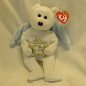 Puede incluir: Un oso de peluche blanco con alas azules y una estrella dorada. El oso tiene ojos negros y una nariz marr&oacute;n. Una etiqueta roja en forma de coraz&oacute;n est&aacute; unida a la oreja. Objeto de colecci&oacute;n.