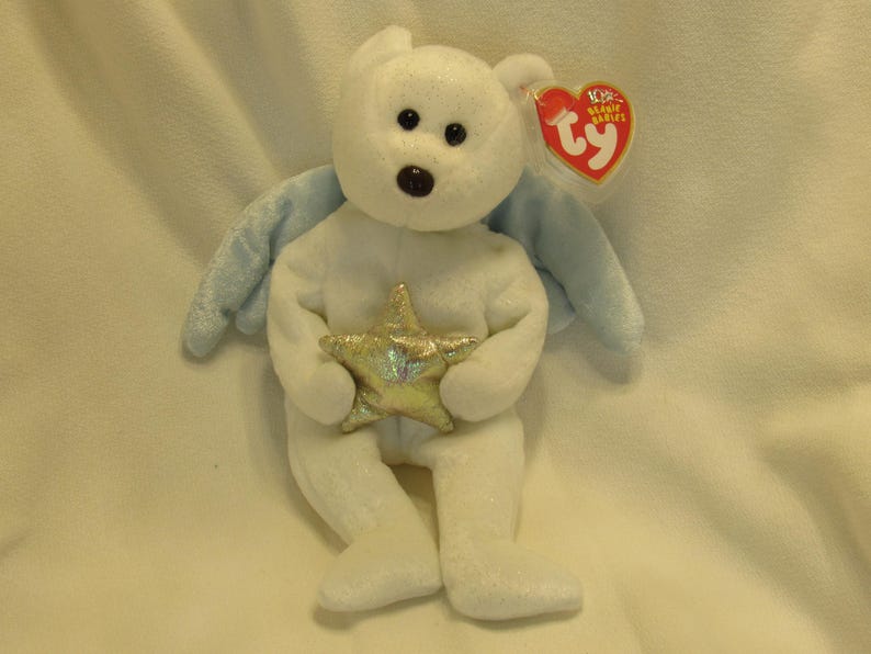 Puede incluir: Un oso de peluche blanco con alas azules y una estrella dorada. El oso tiene ojos negros y una nariz marr&oacute;n. Una etiqueta roja en forma de coraz&oacute;n est&aacute; unida a la oreja. Objeto de colecci&oacute;n.