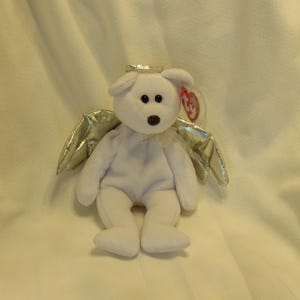 Puede incluir: Un oso de peluche blanco con un halo dorado y alas plateadas. El oso tiene ojos negros y una nariz marr&oacute;n. Una etiqueta roja est&aacute; sujeta a la oreja. El oso est&aacute; sentado sobre una superficie blanca.