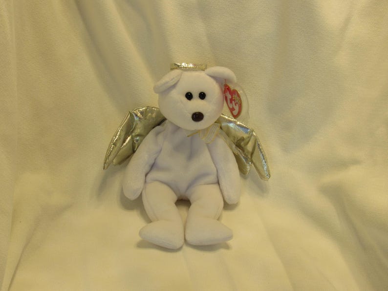 Puede incluir: Un oso de peluche blanco con un halo dorado y alas plateadas. El oso tiene ojos negros y una nariz marr&oacute;n. Una etiqueta roja est&aacute; sujeta a la oreja. El oso est&aacute; sentado sobre una superficie blanca.