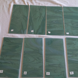 May include: Eight rectangular sheets of green stained glass arranged in two rows of four. Each sheet features a unique marbled pattern with swirls of lighter and darker green tones. The bottom row of four sheets is labeled with white stickers numbered 1 through 4.