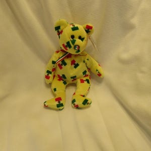 Puede incluir: Un oso de peluche amarillo con detalles rojos, verdes y azules. El oso tiene una cinta roja, blanca y verde alrededor del cuello y una etiqueta en la oreja. El oso est&aacute; sentado sobre una superficie blanca.