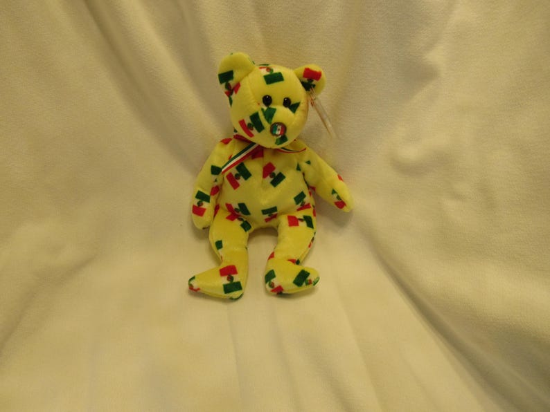 Puede incluir: Un oso de peluche amarillo con detalles rojos, verdes y azules. El oso tiene una cinta roja, blanca y verde alrededor del cuello y una etiqueta en la oreja. El oso est&aacute; sentado sobre una superficie blanca.