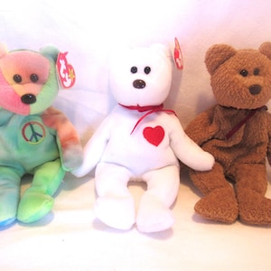 May include: Three plush beanie baby bears. The bear on the left is green, blue, and pink with a peace sign on its chest. The bear in the middle is white with a red heart on its chest. The bear on the right is brown.