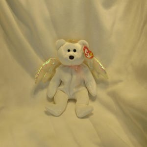 Puede incluir: Un oso de peluche blanco con alas de &aacute;ngel iridiscentes y un halo dorado. El oso tiene una nariz negra, una etiqueta roja y una cinta rosa alrededor del cuello. Un peluche coleccionable.
