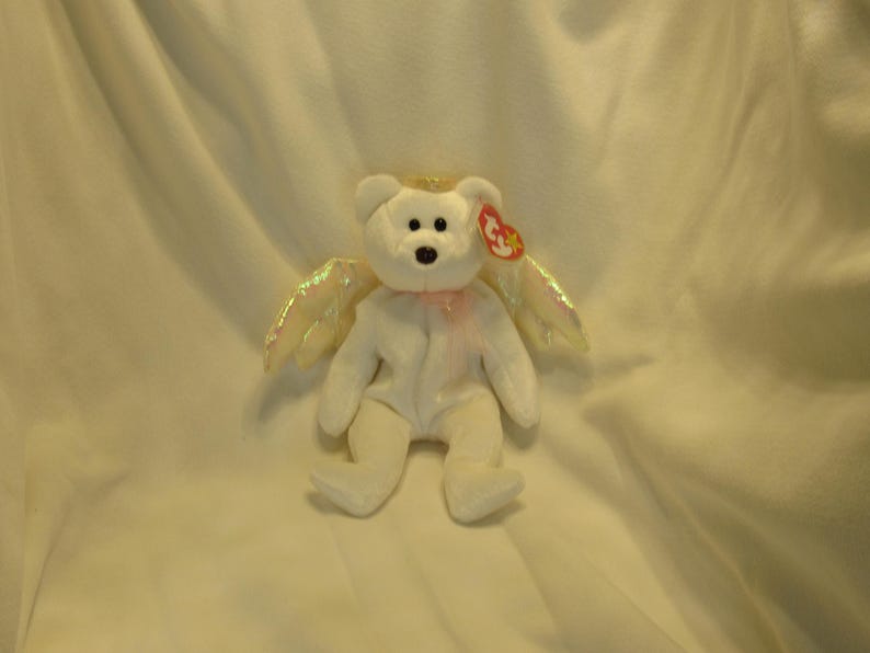 Puede incluir: Un oso de peluche blanco con alas de &aacute;ngel iridiscentes y un halo dorado. El oso tiene una nariz negra, una etiqueta roja y una cinta rosa alrededor del cuello. Un peluche coleccionable.