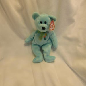 Puede incluir: Un oso de peluche azul claro con una cinta verde y un dise&ntilde;o floral. El oso tiene una etiqueta roja. El oso mide aproximadamente 20 cm de altura. Un juguete infantil cl&aacute;sico.
