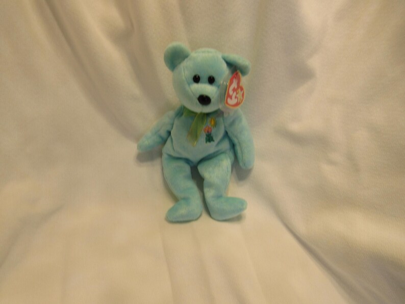 Puede incluir: Un oso de peluche azul claro con una cinta verde y un dise&ntilde;o floral. El oso tiene una etiqueta roja. El oso mide aproximadamente 20 cm de altura. Un juguete infantil cl&aacute;sico.