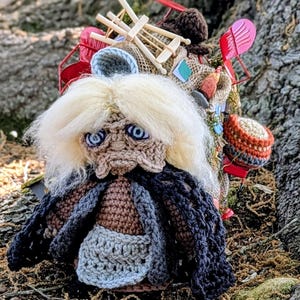 May include: A handmade crochet doll with a whimsical design. The doll has a light blonde yarn hair, blue eyes, and a dark jacket. It is sitting in front of a tree trunk, with moss and twigs in the foreground. A backpack is behind the doll.