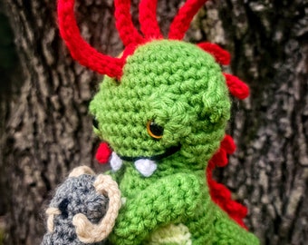 May include: A green crocheted dinosaur with red spikes and white teeth holds a gray crocheted animal in its arms. The dinosaur is sitting on a wooden surface.
