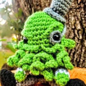 Może przedstawiać: Zielony i szary ośmiornica na szydełku z czarną łatką na oku i białymi akcentami siedzący na drewnianej powierzchni.