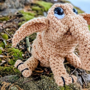 Op de afbeelding: Een beige gehaakt wezen met grote zwarte en witte ogen, lange oren en kleine zwarte accenten op de voeten. Het handgemaakte pluche speelgoed zit op een mosachtige ondergrond en toont zijn gedetailleerde steken en grillige ontwerp.
