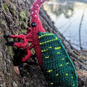 May include: A crocheted green and red bug with yellow and white accents. The bug has a long snout and is perched on a tree trunk.