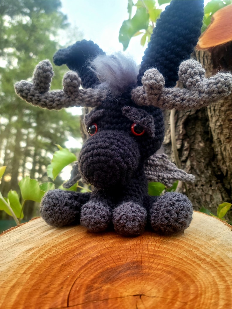 May include: A black and gray crocheted moose with red eyes and antlers. The moose is sitting on a wooden log.