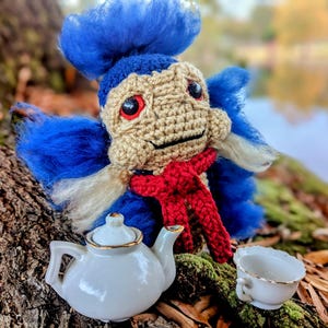 May include: A crocheted blue and white creature with a red scarf sits on a mossy surface with a white teapot and a white teacup.