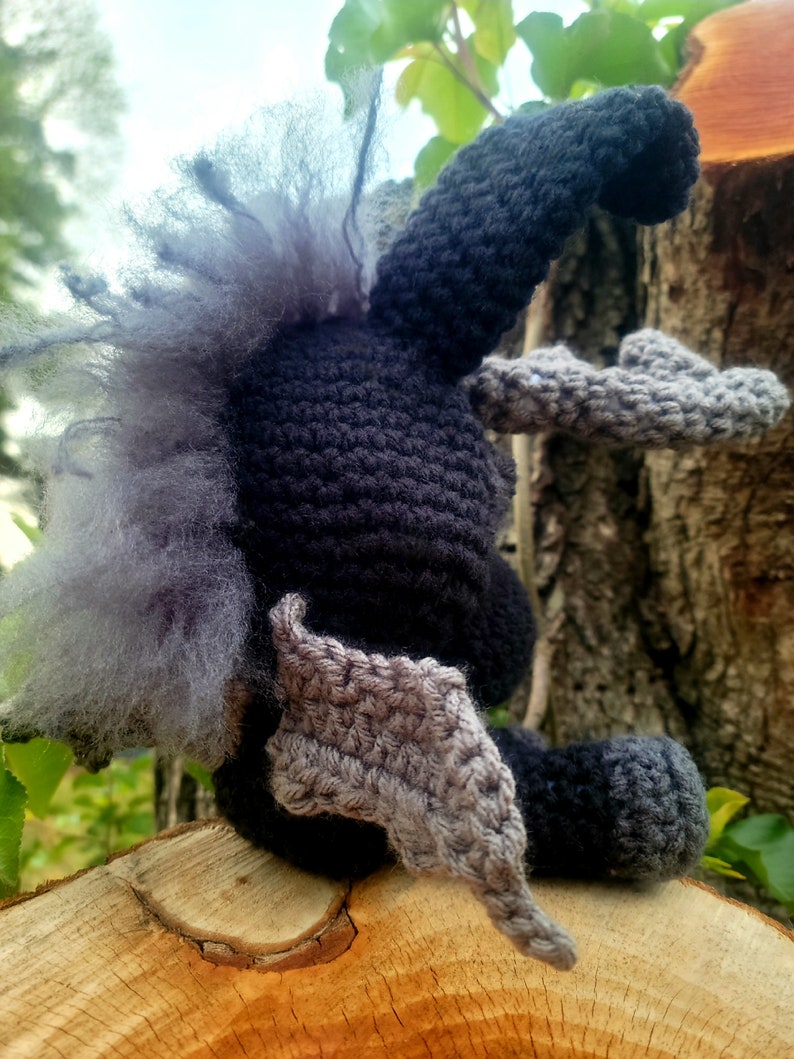 May include: A crocheted plush toy with gray and black fur, wings, and a tail. The toy is sitting on a wooden surface.