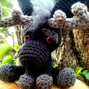 May include: A crocheted moose plush toy with black and gray fur, red eyes, and large antlers. The moose is sitting on a wooden surface.