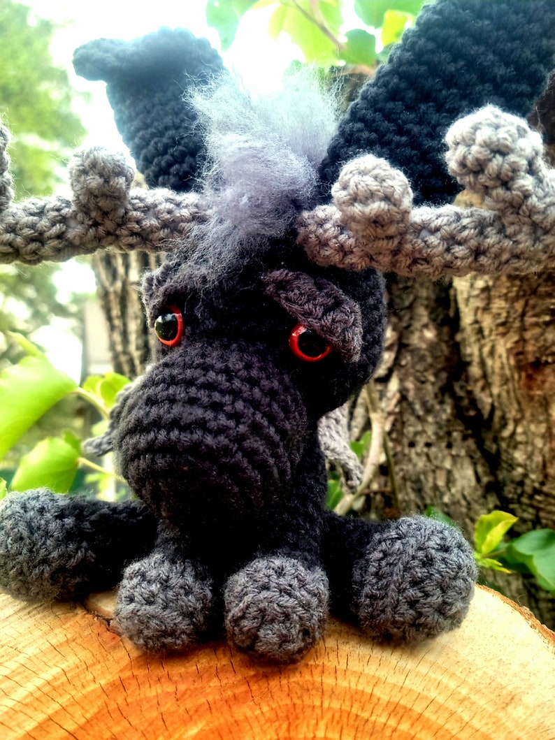 May include: A crocheted moose plush toy with black and gray fur, red eyes, and large antlers. The moose is sitting on a wooden surface.