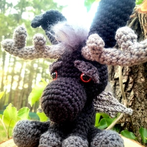 May include: A crocheted black and gray moose with red eyes and a furry mane. The moose has antlers and wings. It is sitting on a wooden surface.
