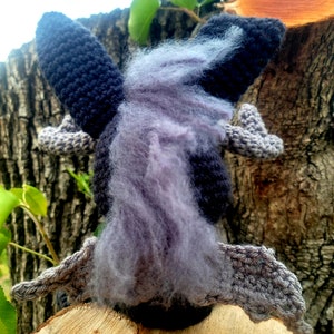 May include: A crocheted plush toy shaped like a bat with gray wings and a black body. The bat has a long, fluffy, purple tail.