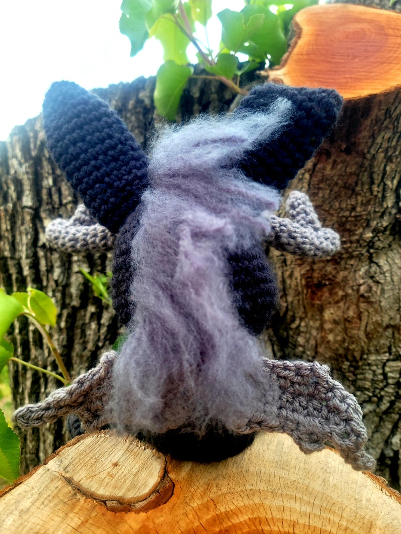 May include: A crocheted plush toy shaped like a bat with gray wings and a black body. The bat has a long, fluffy, purple tail.