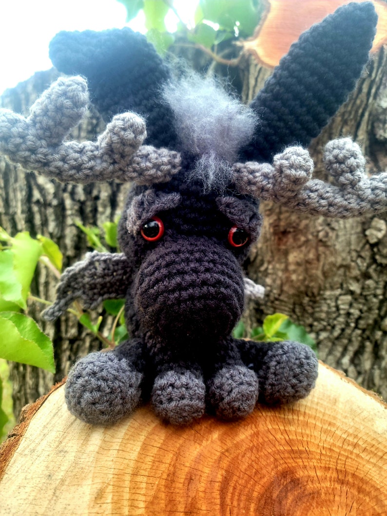 May include: A crocheted black moose with red eyes and gray antlers. The moose is sitting on a wooden log.