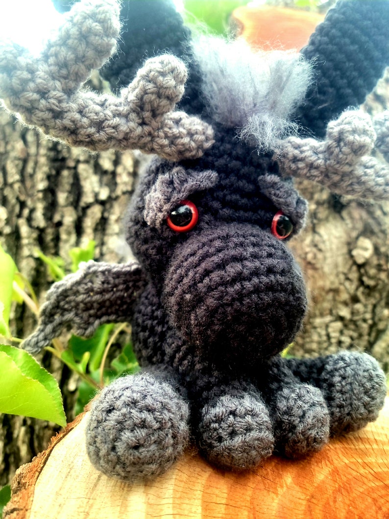 May include: A crocheted black moose with gray antlers and red eyes. The moose is sitting on a wooden surface.
