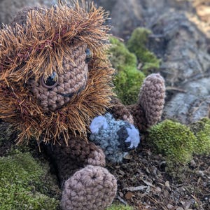 May include: A brown crocheted plush toy with a furry brown and orange mane, holding a gray crocheted plush toy. The toys are sitting on a bed of green moss.