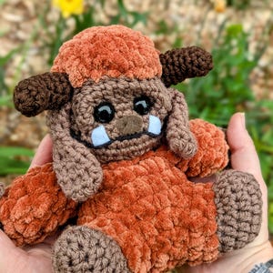 Op de afbeelding: Een gehaakt pluche speelgoed in de vorm van een bruin en oranje monster met grote ogen en een brede grijns. Het speelgoed zit in de palm van een hand.