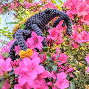 May include: A handmade crochet insect with a long snout, in shades of gray, yellow, and brown, perched among vibrant pink azalea flowers. The amigurumi-style creature has large black eyes and detailed legs, creating a whimsical garden scene.