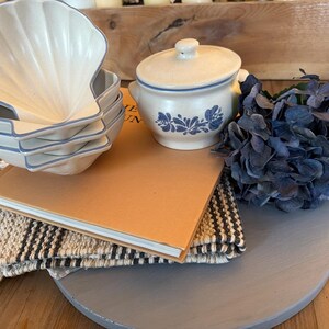 May include: A stack of four off-white ceramic bowls shaped like seashells with blue rims, a matching off-white lidded sugar bowl with blue floral patterns, a tan hardcover book, a woven beige and black textile, and a cluster of dark blue flowers.
