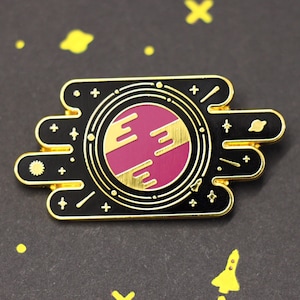Puede incluir: Pin de esmalte dorado con un diseño negro y dorado. El pin presenta un planeta rosa con anillos dorados y un fondo negro. El pin está rodeado de pequeñas estrellas doradas y otros detalles con temática espacial.