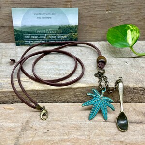 May include: A brown leather necklace with a bronze clasp and a turquoise maple leaf pendant. A small bronze spoon charm hangs next to the leaf. A business card with the text "Forest Kandi" is in the background.