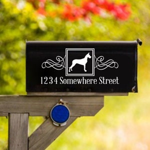 May include: A black mailbox with a white dog silhouette and the address "1234 Somewhere Street" on a wooden post. The mailbox is surrounded by green foliage and a dirt road.
