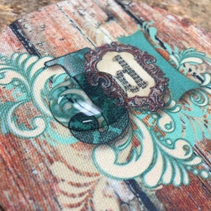 May include: A round coaster with a wood grain pattern and a teal blue and white floral design. The center of the coaster features a teal blue and white floral design with a gold frame and the number "1" in the center.