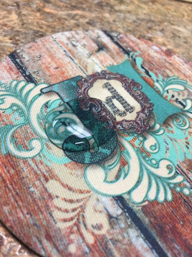 May include: A round coaster with a wood grain pattern and a teal blue and white floral design. The center of the coaster features a teal blue and white floral design with a gold frame and the number "1" in the center.