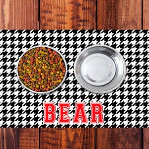 May include: Black and white houndstooth patterned pet food mat with two stainless steel bowls, one filled with dry pet food. The mat has the word "BEAR" in red letters.