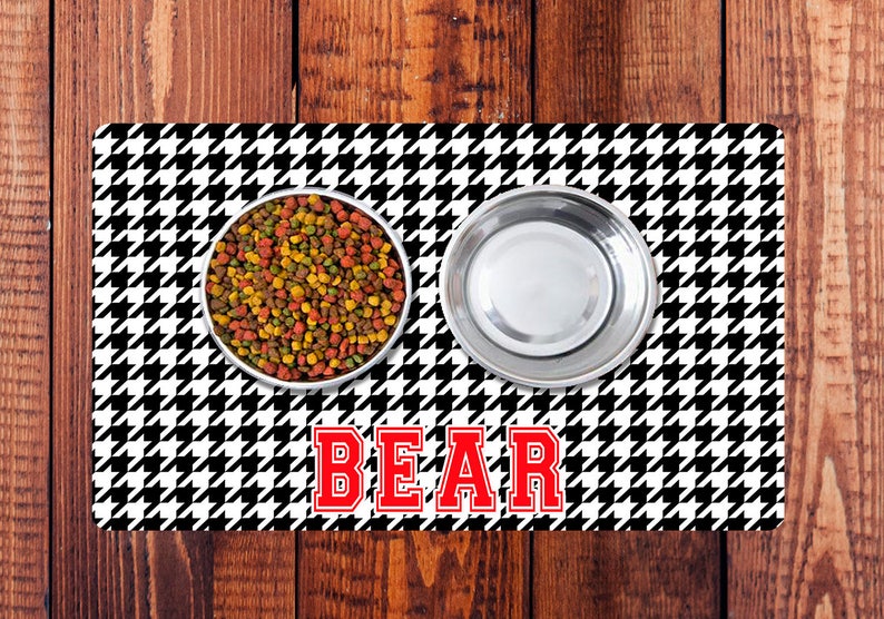 May include: Black and white houndstooth patterned pet food mat with two stainless steel bowls, one filled with dry pet food. The mat has the word "BEAR" in red letters.