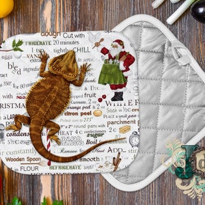 Puede incluir: Manopla de cocina blanca con textura acolchada. La parte delantera presenta un lagarto barbudo marrón con un bastón de caramelo rojo y un fondo blanco con una receta navideña impresa en él. La receta incluye instrucciones para hacer un fudge blanco.