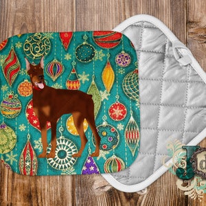 May include: A teal blue pot holder with a brown Doberman Pinscher dog standing in front of a Christmas ornament pattern. The ornaments are red, green, gold, and white. The pot holder is white on the back.