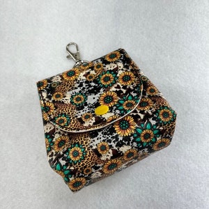 May include: A small, square-shaped pouch with a zipper closure and a metal clip. The pouch is made of a fabric with a white background and a pattern of brown leopard spots and yellow sunflowers with green centers. The pouch has a yellow button on the front.