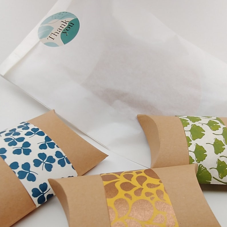 May include: Three small, brown, pillow-shaped boxes with different patterned paper designs. The boxes are on a white surface. One box has a blue and white clover pattern, another has a gold and brown floral pattern, and the third has a green and white ginkgo leaf pattern.